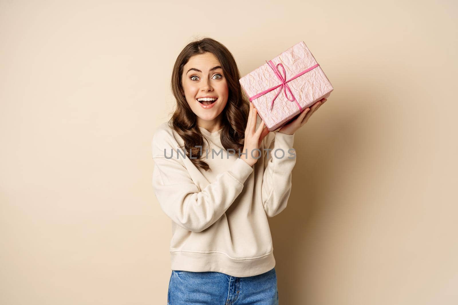 Excited cute girl shaking box with gift, guessing whats inside present, standing happy against beige background.