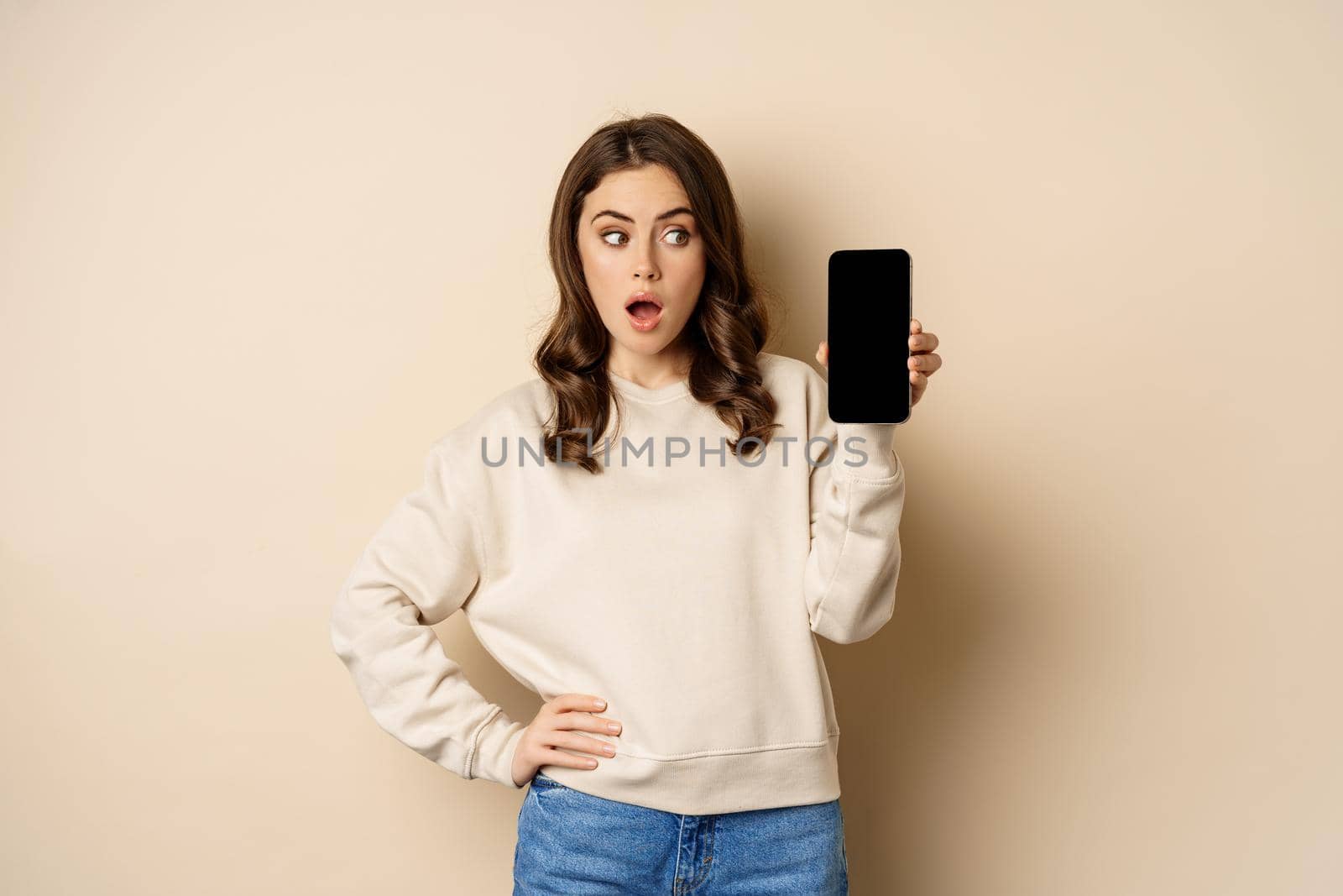 Woman showing smartphone screen and looking surprised at mobile phone, standing in sweater over beige background by Benzoix