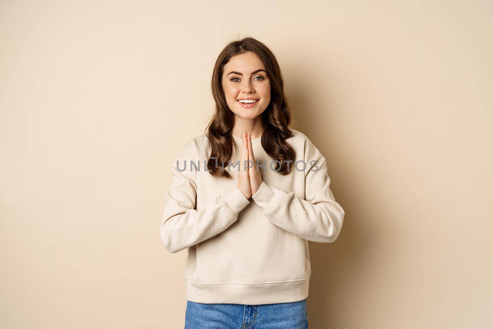 Thank you. Smiling happy woman showing pray, namaste gesture, thanking, standing pleased and grateful over beige background by Benzoix