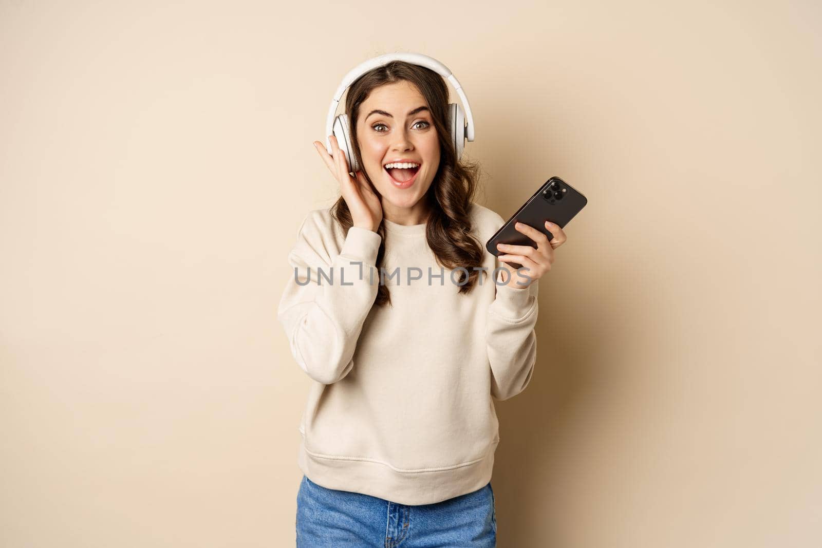 Smiling happy woman listening music in headphones on smartphone, laughing excited, standing over beige background.