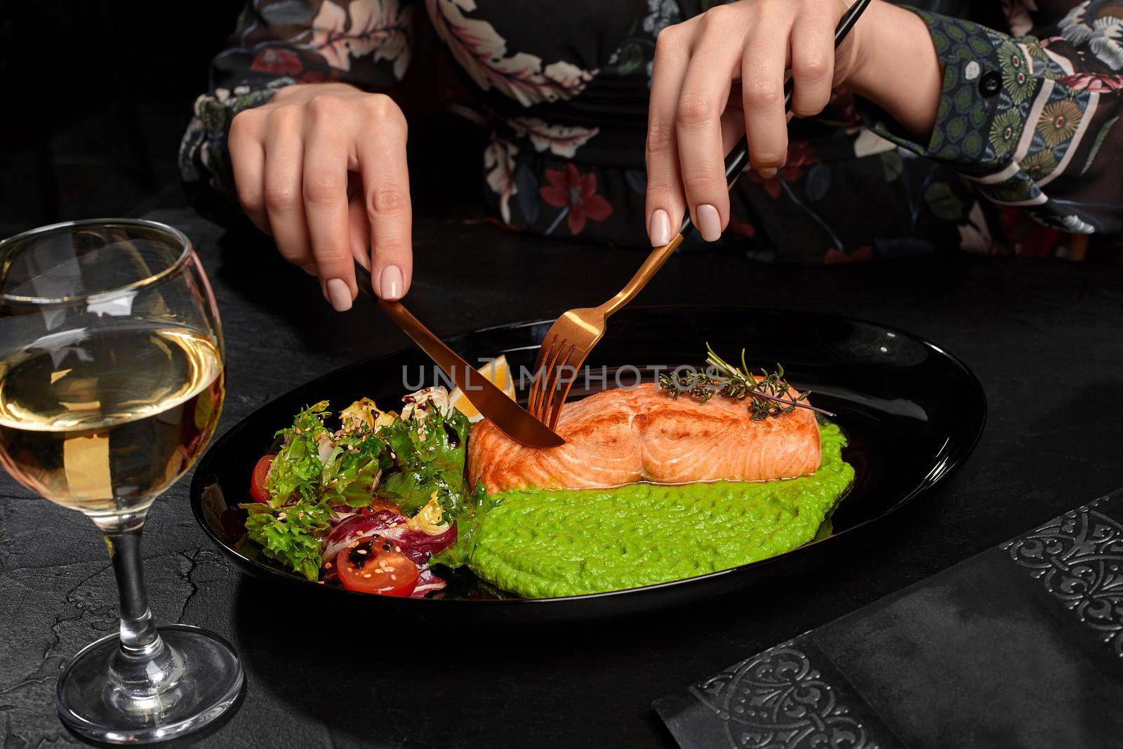 Female hands cutting grilled salmon steak served with pea puree, fresh salad, lemon and glass of wine by nazarovsergey