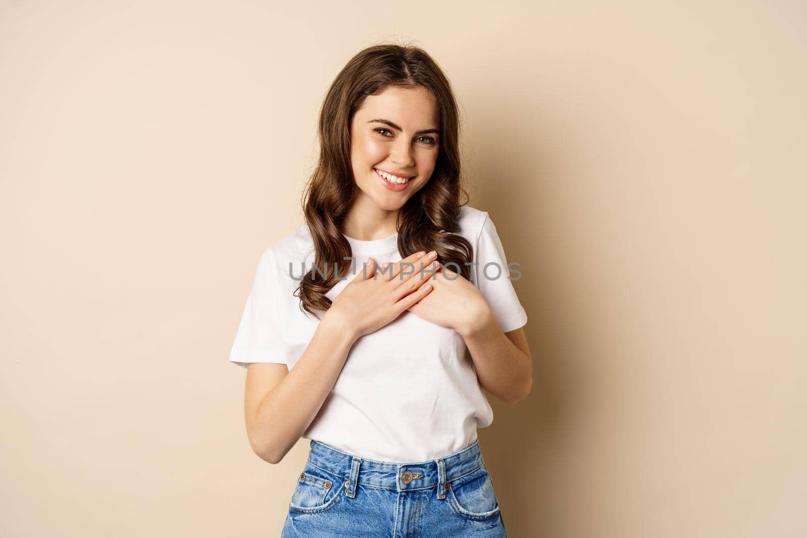 Portrait of young caucasian woman hold hands on heart and smiling, daydreaming, thinking of smth heartwarming, posing against beige background by Benzoix