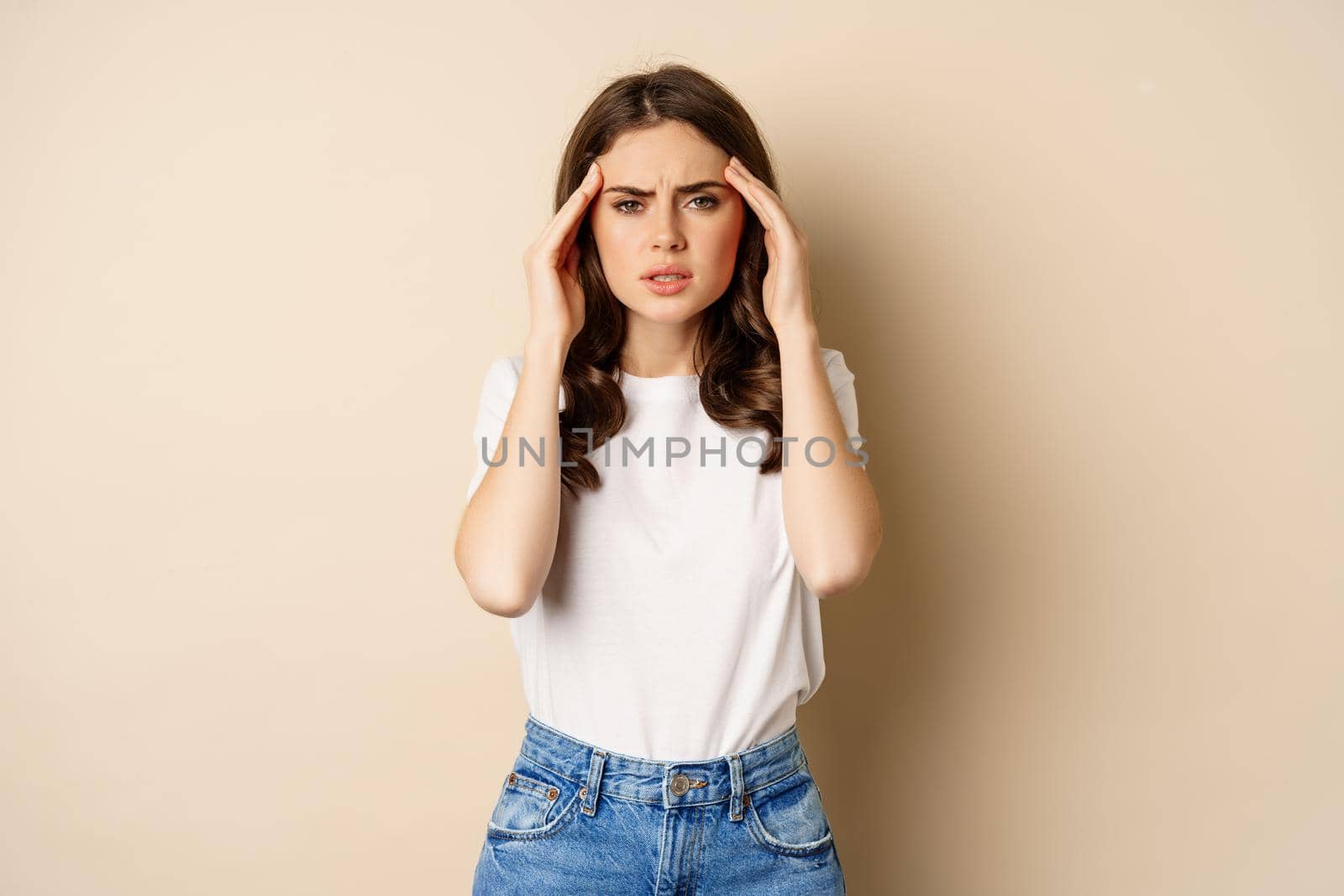 Health and women concept. Portrait of girl feeling sick, touching head temples, feeling headache, migraine, standing over beige background.