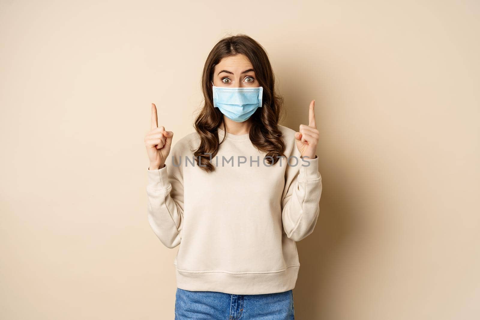 Happy young woman in medical mask from covid-19, pointing fingers up, showing promo or copy space, standing over beige background.