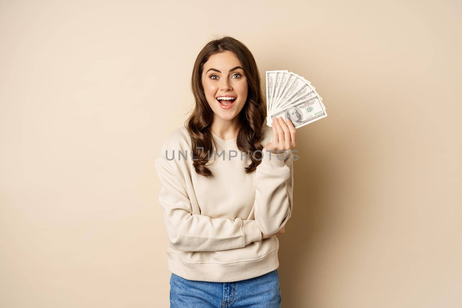 Happy woman showing money, cash and smiling pleased, concept of microcredit and shopping, standing over beige background by Benzoix