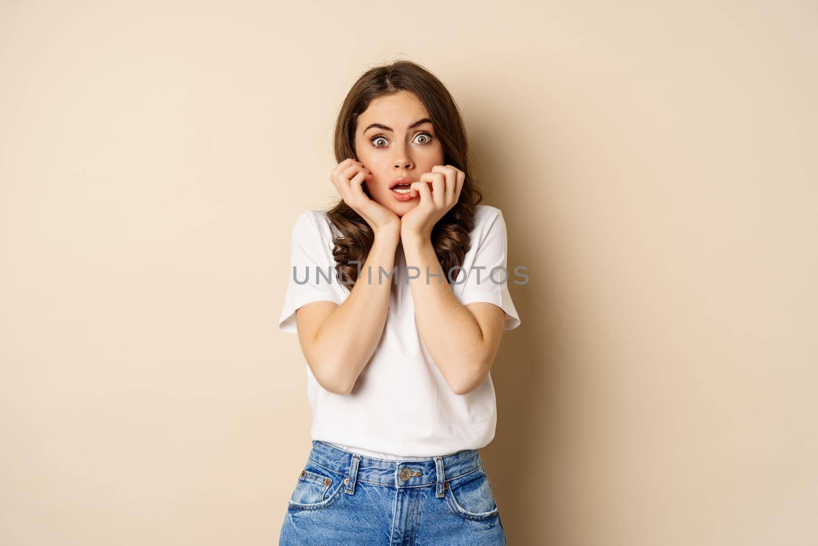 Scared and shocked woman gasping, looking startled speechless, wearing t-shirt, standing over beige background.