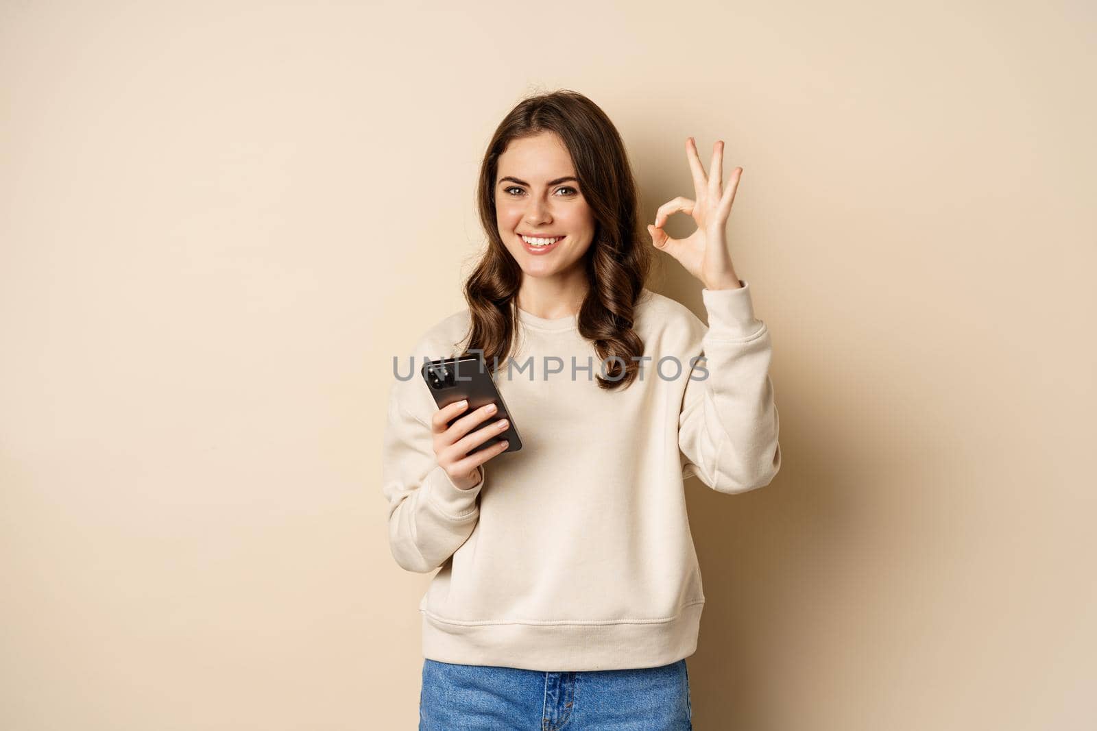 Beautiful woman holding cellphone, mobile phone and okay sign, recommending application, shopping app, standing over beige background by Benzoix