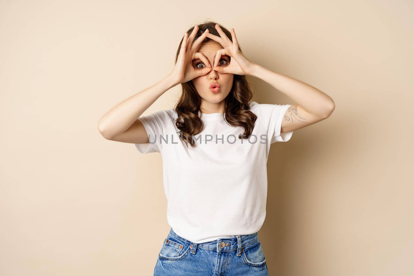 Funny brunette woman having fun, showing finger glasses gesture and fool around, posing over beige background.
