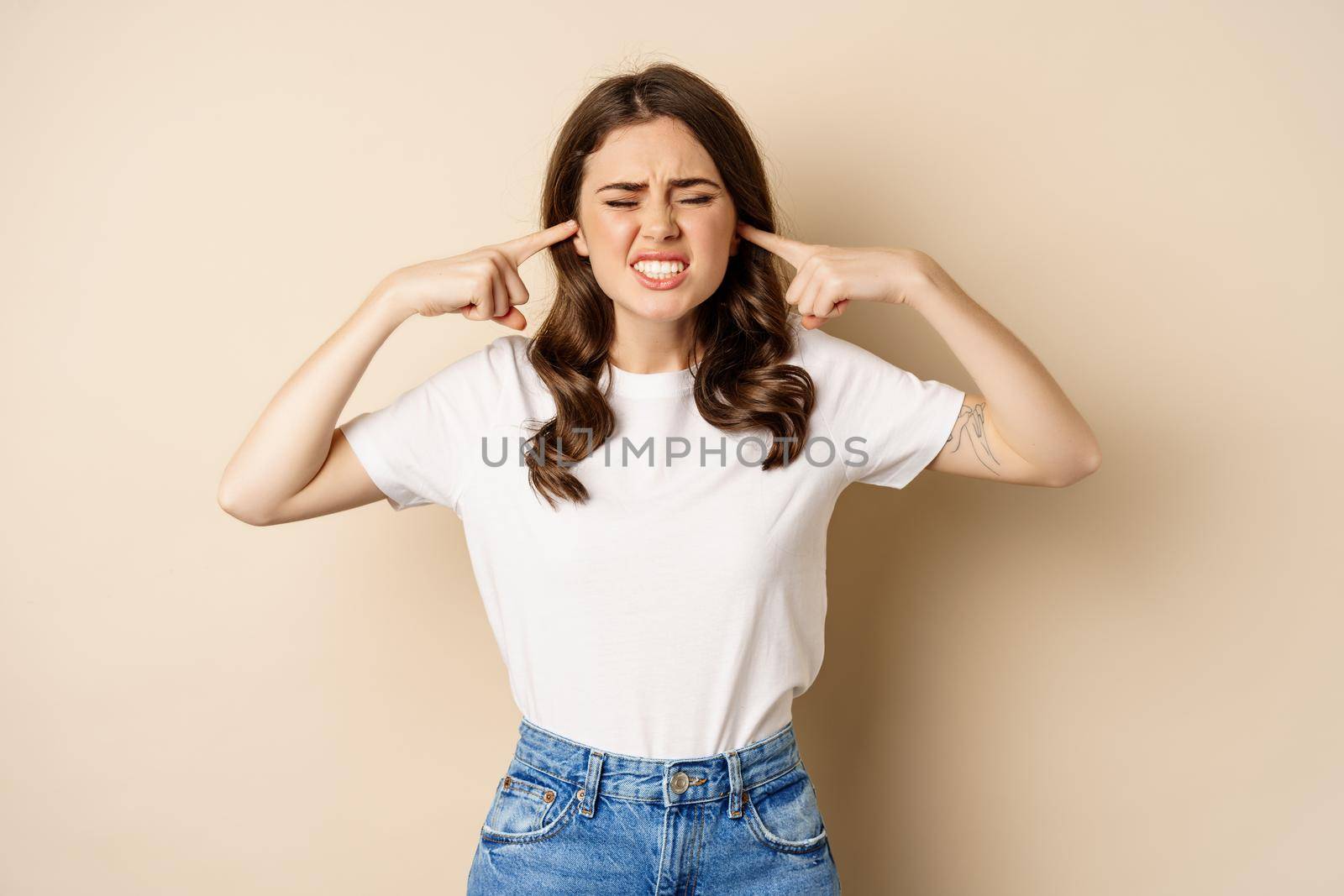 Annoyed young woman shut ears from loud noise, feeling discomfort and complaining, standing against beige background.