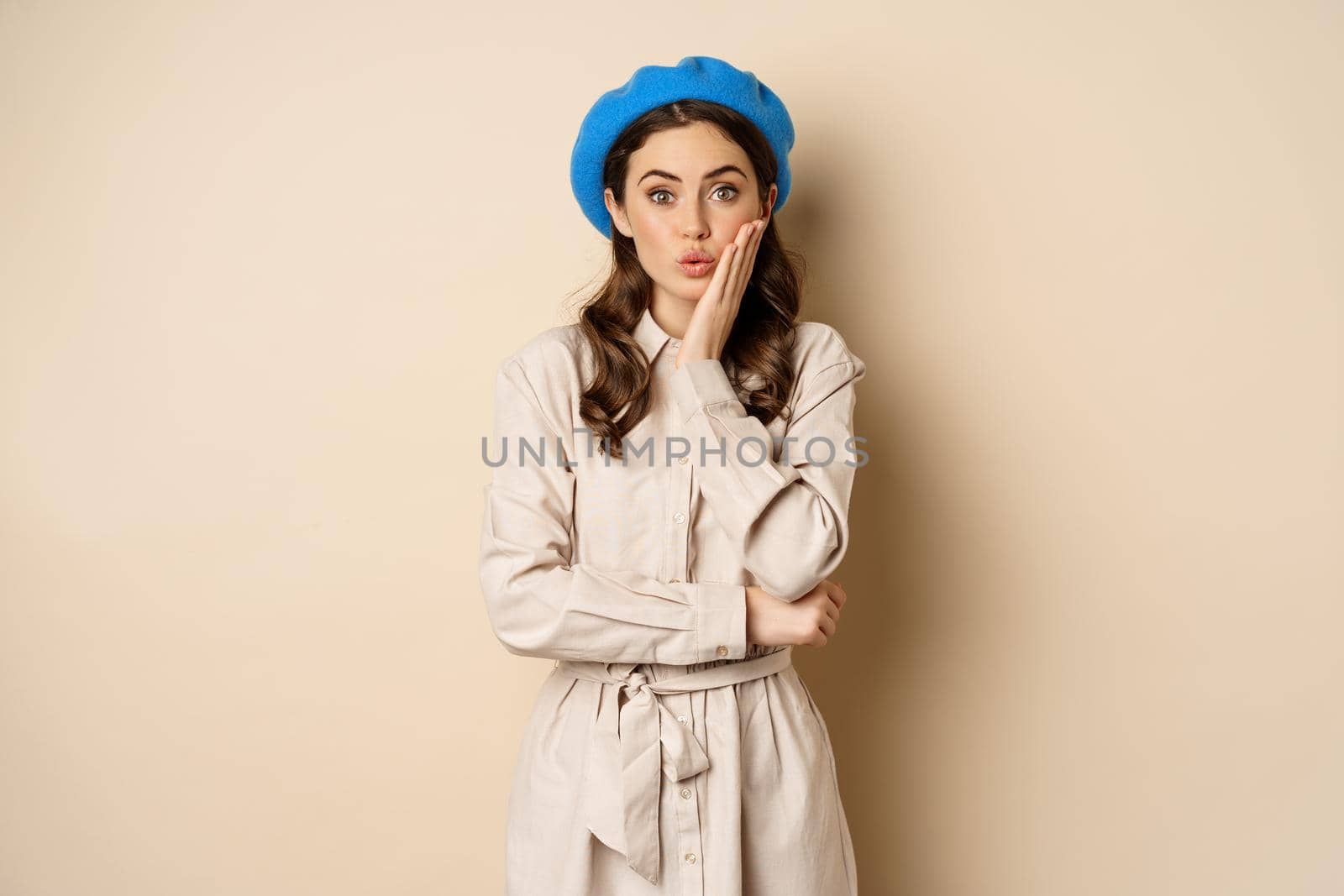 Image of stylsh beautiful woman looking surprised, shocked reaction at camera, posing in trench coat against beige background.