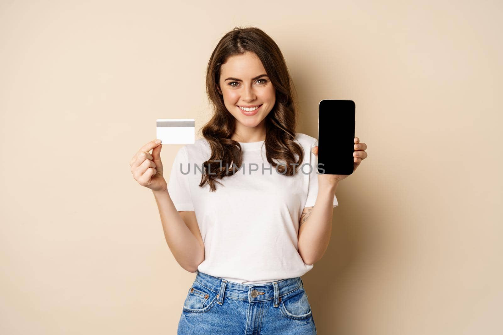 Online shopping and people concept. Young beautiful woman looking happy, showing credit card discount and mobile phone screen, standing over beige background.