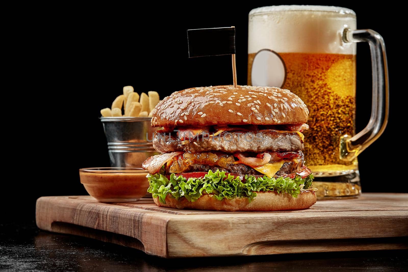 Hamburger with two juicy patties, fried bacon, cheese, caramelized onions, fresh tomatoes and lettuce in fluffy sesame bun served on wooden board with glass of light beer, french fries and aioli sauce