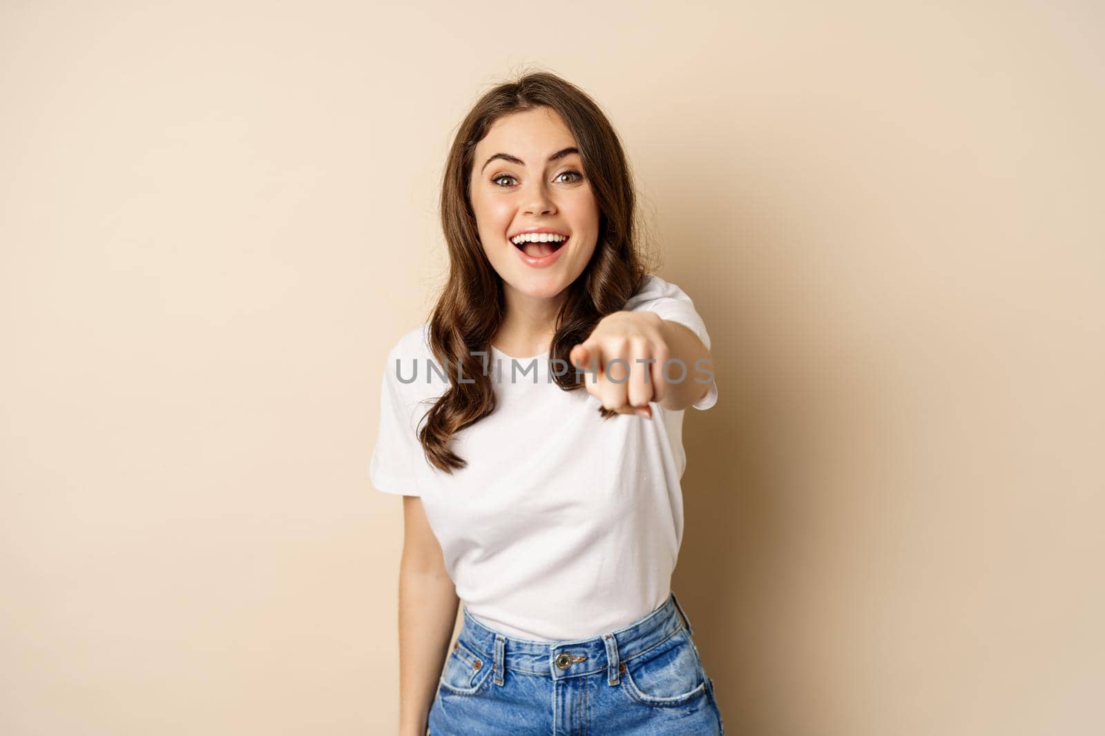 Its you, congrats. Smiling beautiful woman pointing finger at camera, congratulating, praising you, standing over beige background.