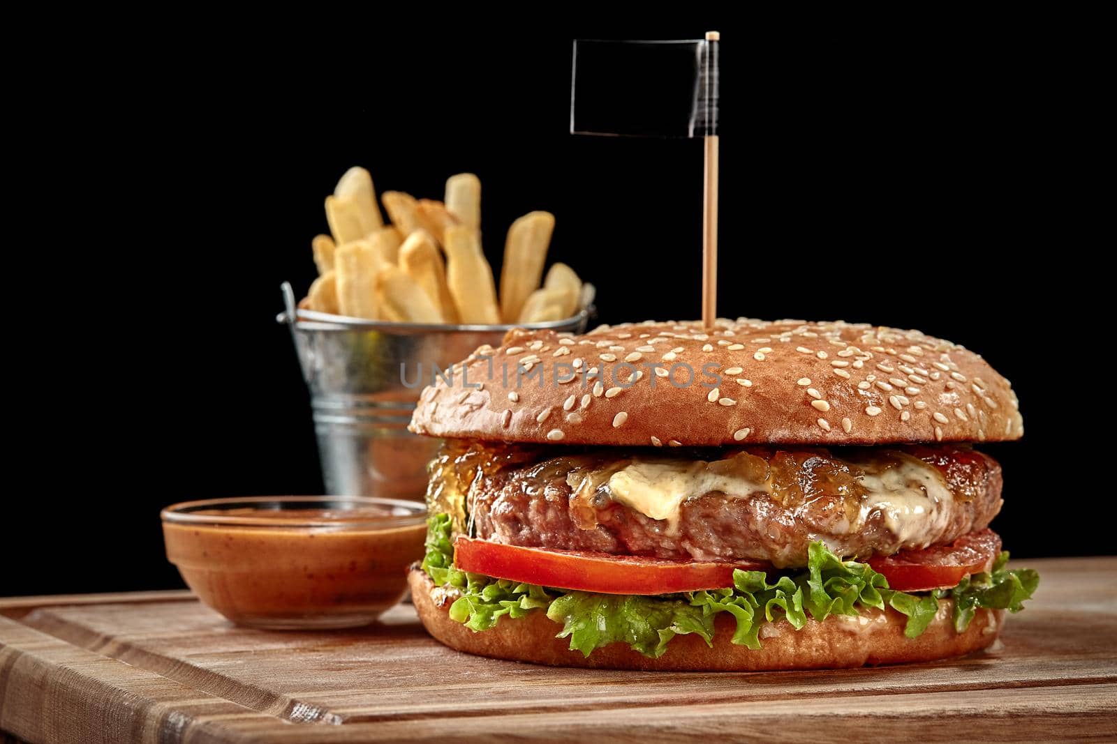 Big hearty hamburger with juicy beef patty, creamy blue cheese, caramelized onion, fresh tomatoes and lettuce in fluffy bun with sesame served with crispy french fries and spicy sauce on wooden board