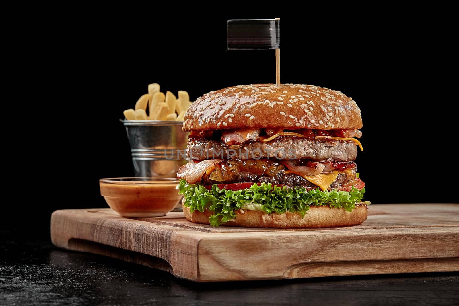 Delicious burger with two juicy pork and beef patties, fried bacon, cheese slices, caramelized onions, fresh tomatoes and greens in fluffy sesame bun served with fries and aioli sauce on wooden board