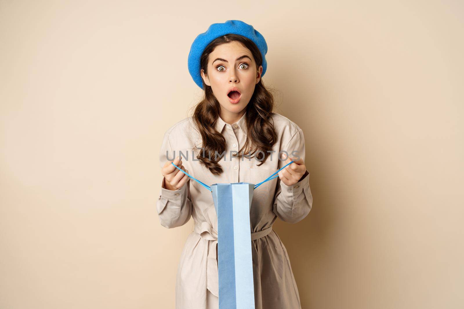 Image of girl looking surprised after opening gift bag, looking at present amazed, standing in stylish outfit over beige background.