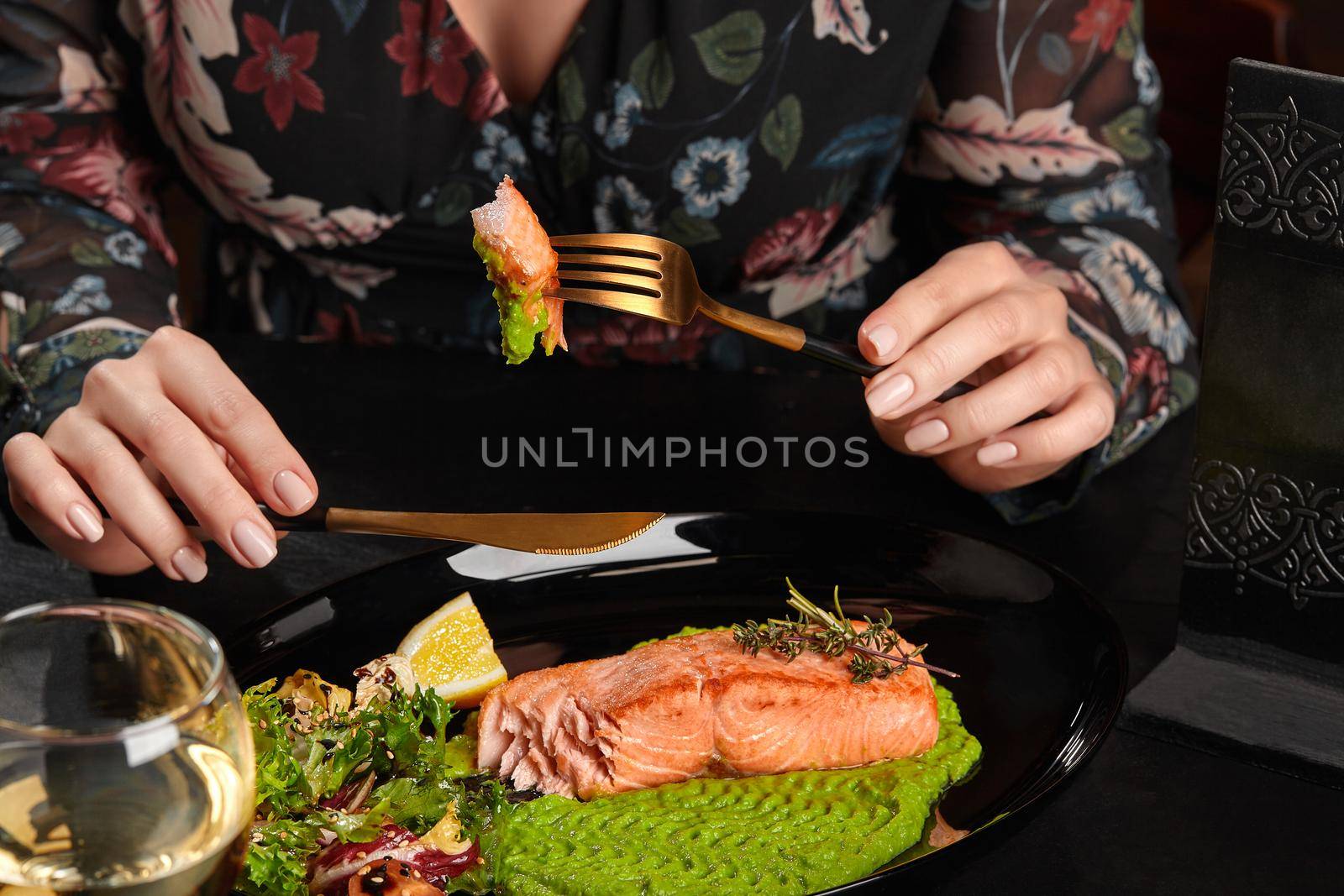 Hands of young woman holding knife and fork with piece of appetizing baked trout steak served with mashed green peas, fresh salad, lemon slice and aromatic herbs