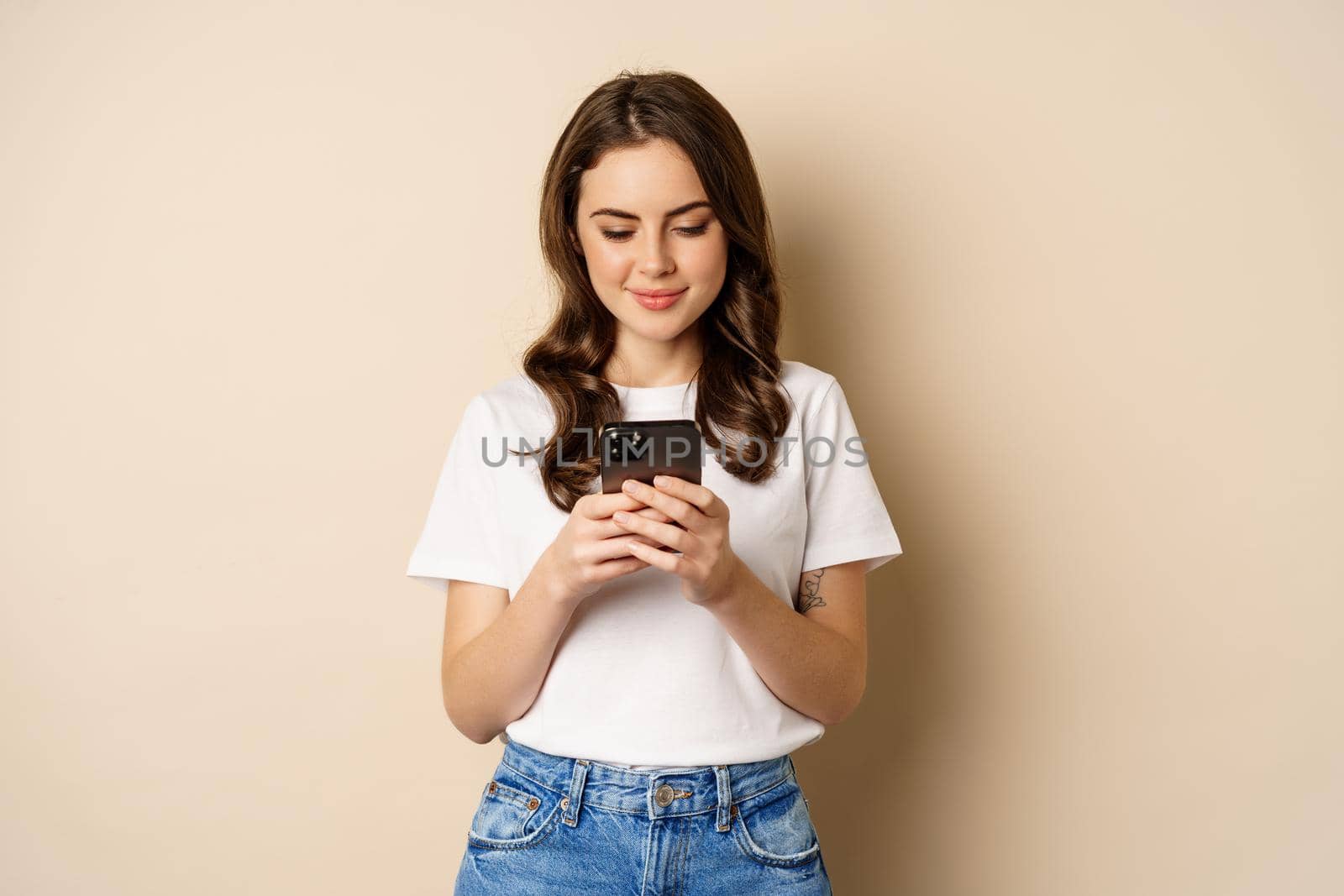 Young modern girl chatting on app, using smartphone app and smiling, standing over beige background by Benzoix