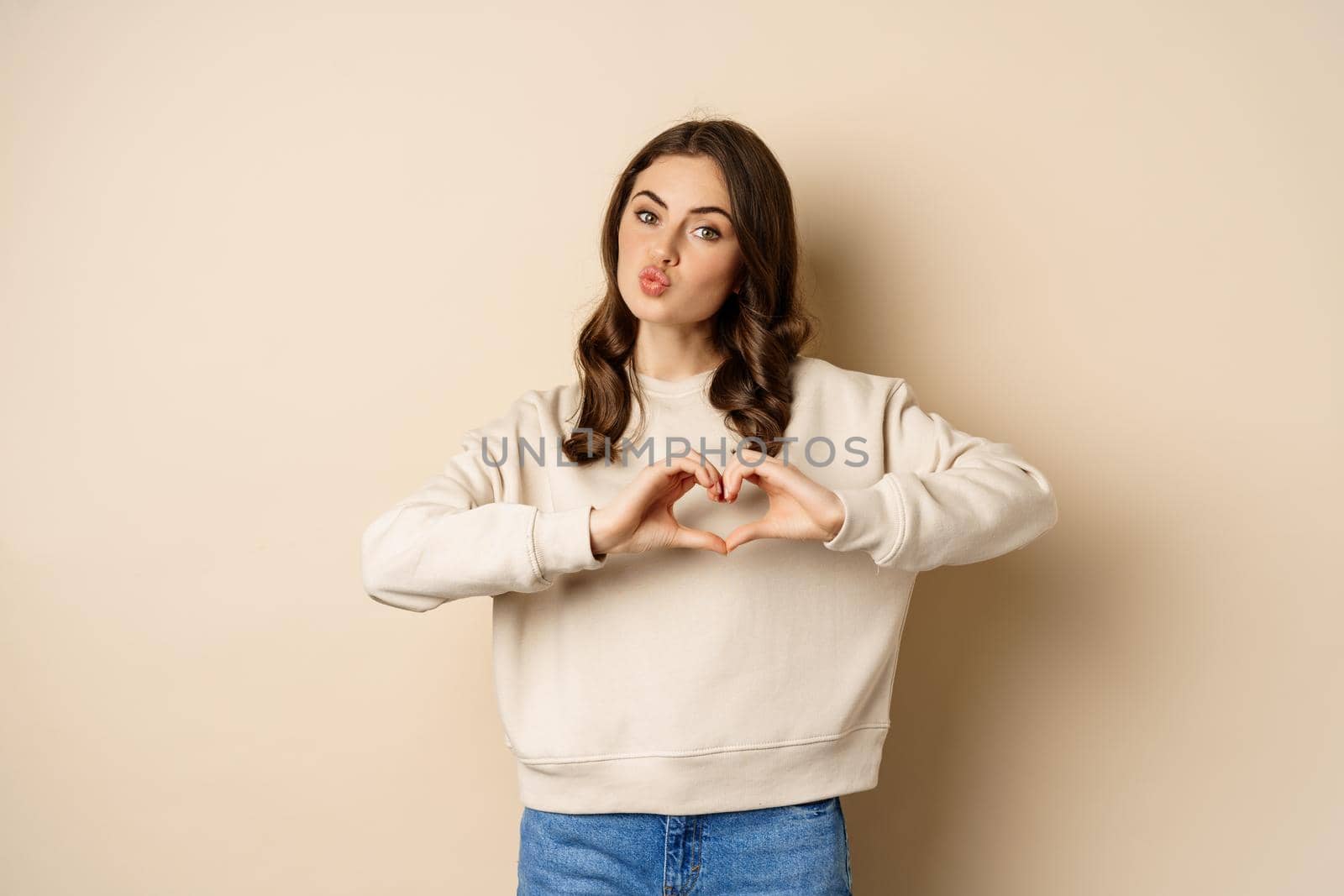 Beautiful, romantic brunette woman showing heart, love sign, care for you, smiling at camera, standing over beige background by Benzoix