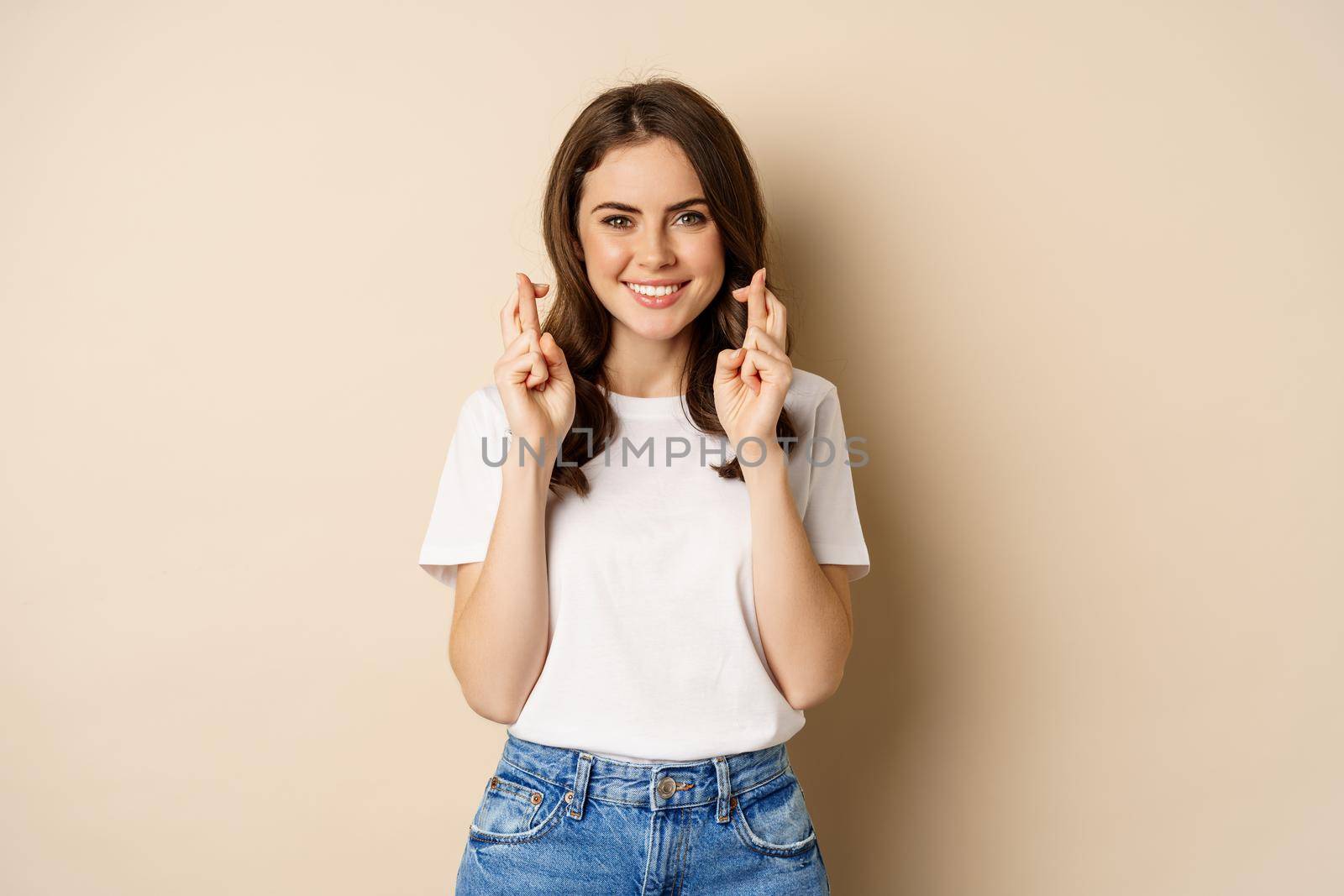 Hopeful woman praying, cross fingers for good luck, making wish, standing over beige background.