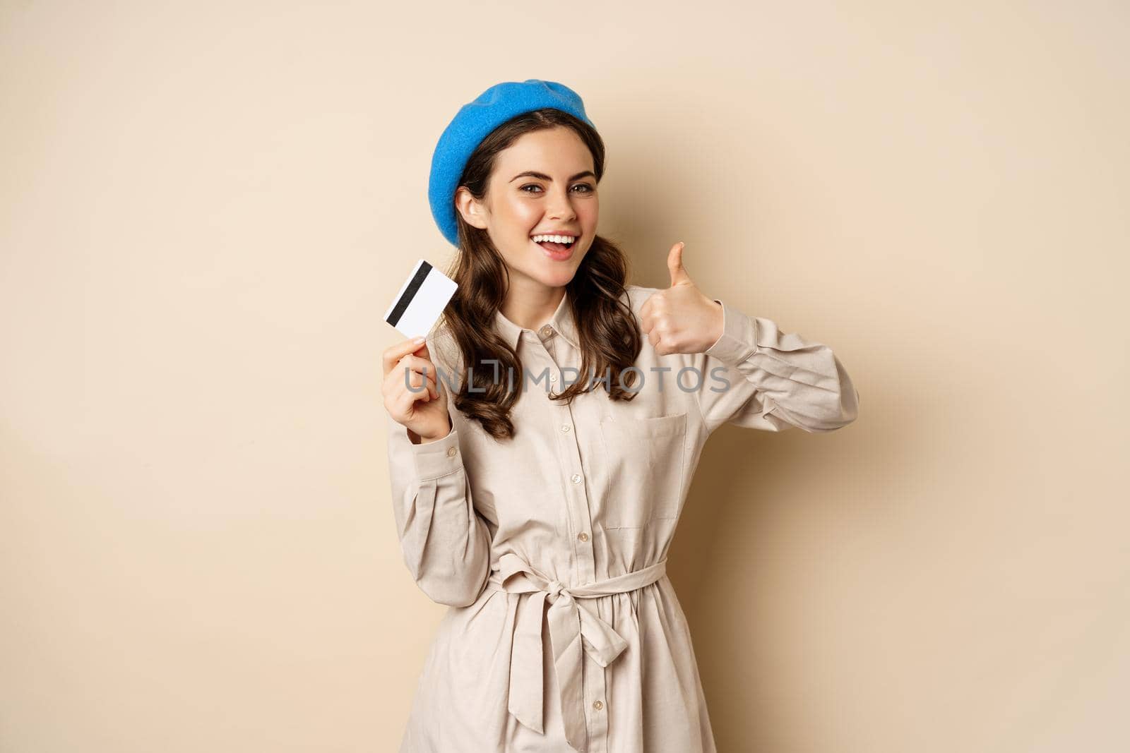 Concept of shopping and sale. Happy young woman in stylish clothes, showing credit card store discount and thumbs up, recommending, standing over beige background by Benzoix