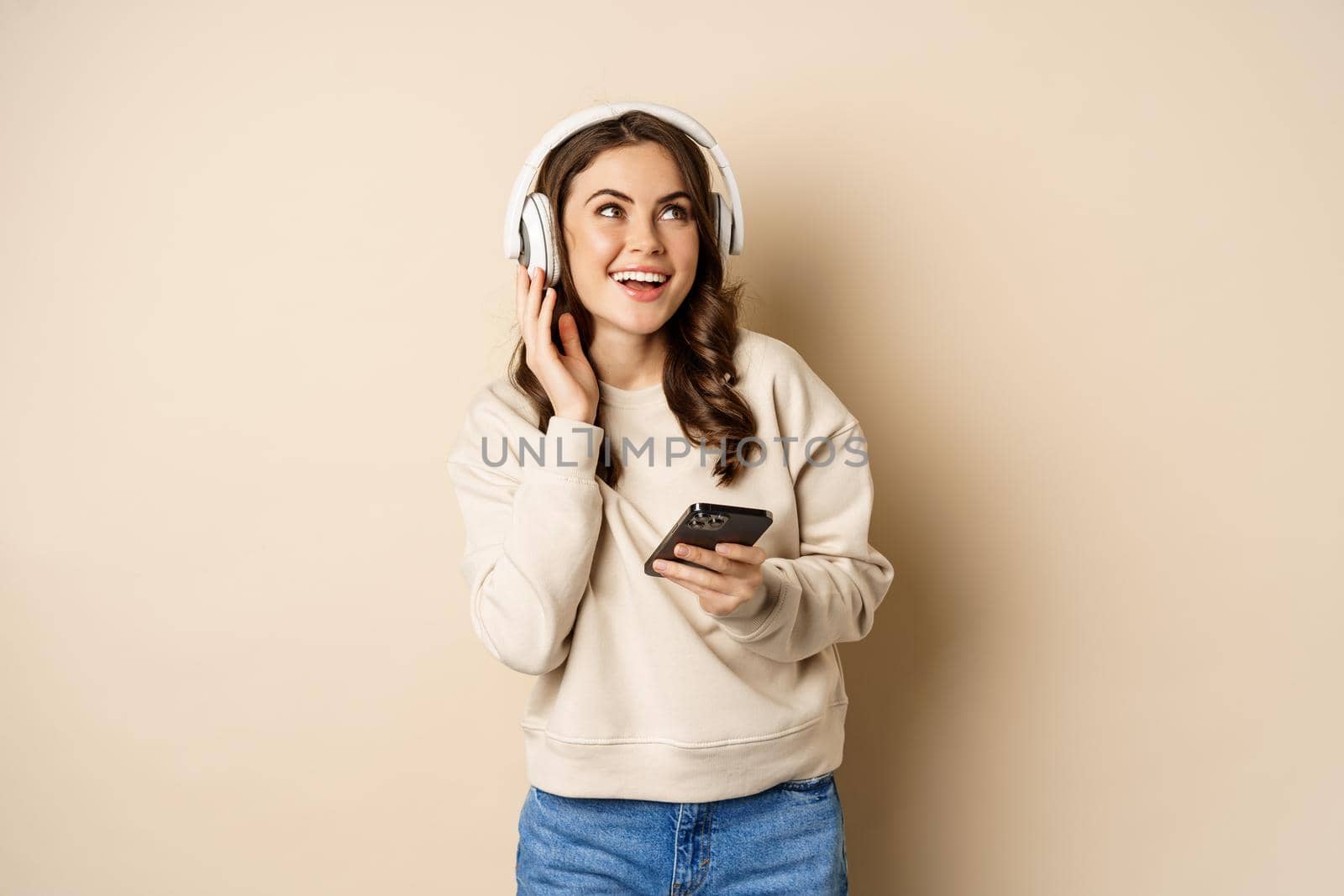 Happy caucasian woman listening music in headphones on smartphone app, dancing and having fun, standing over beige background.