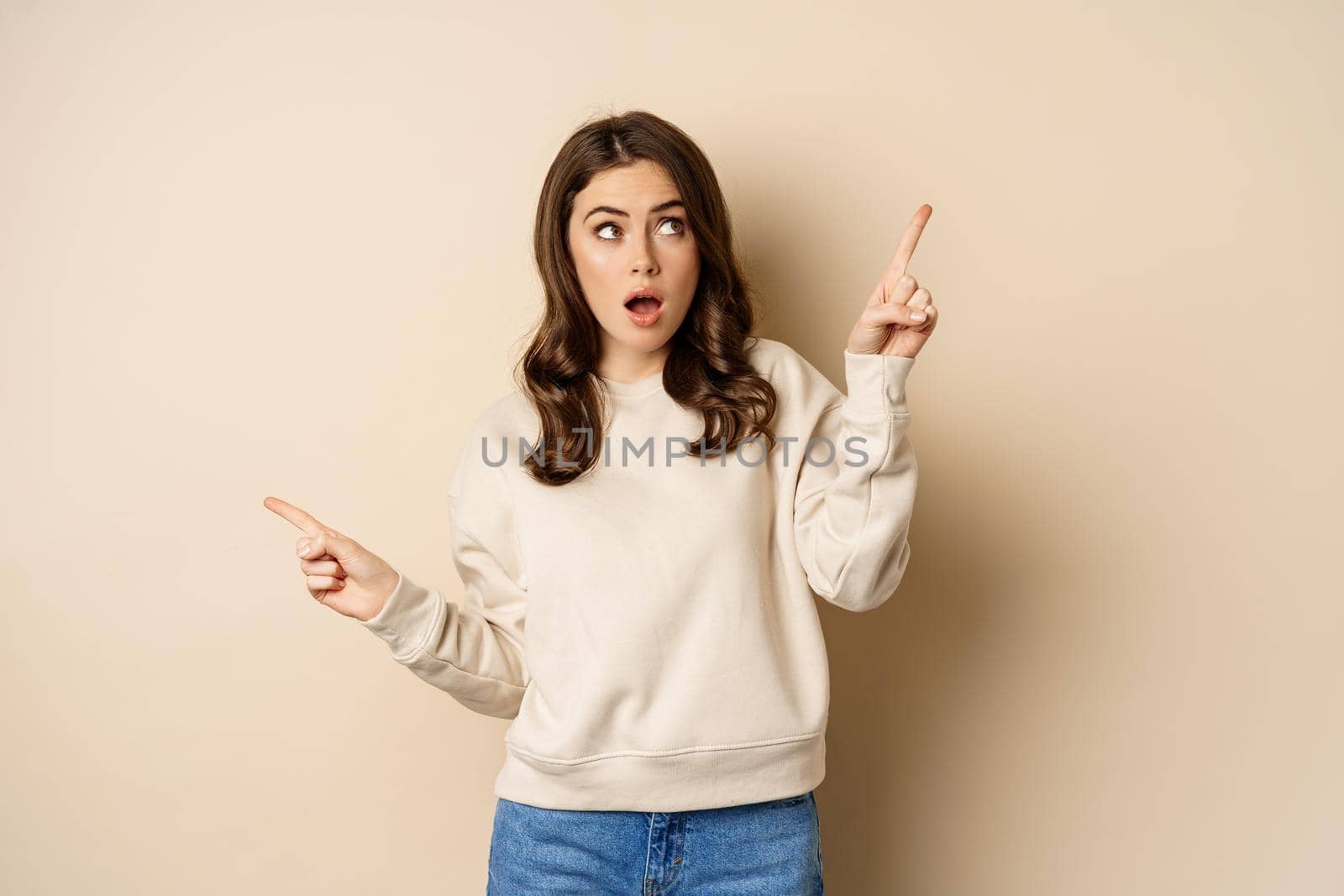 Confused girl shopper pointing sideways, making choice, decision between two variants, beige background. Copy space