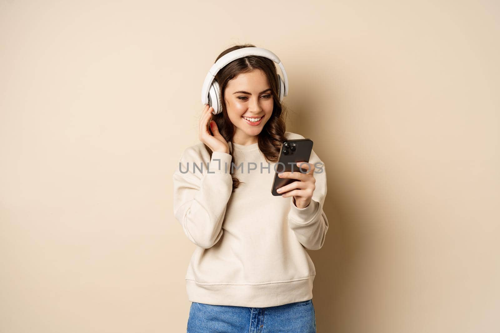 Beautiful young woman looking at video on smartphone, listening music in headphones, standing in casual clothes over beige background by Benzoix