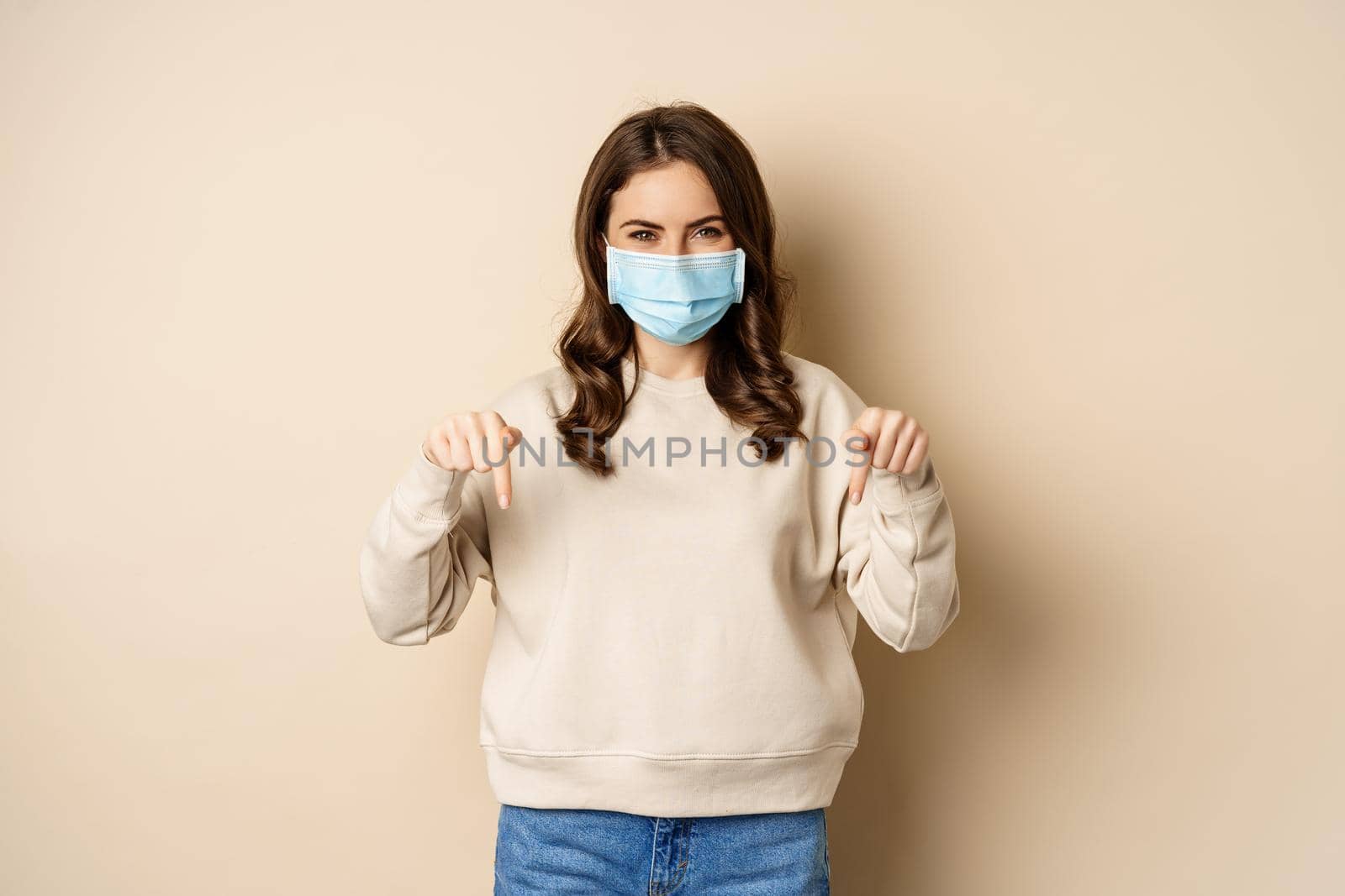 Cheerful cute woman in medical face mask, pointing fingers down, showing advertisement, using protection from covid-19, beige background.