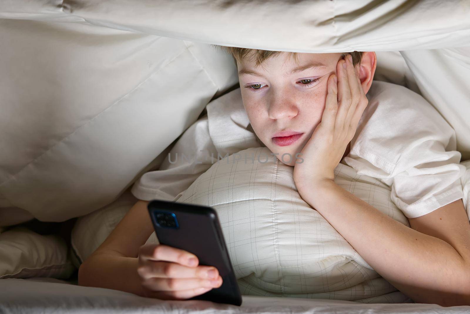 Boy under the blanket at night in his bed communicates on Internet. Child gadget addiction and insomnia. social media addiction by PhotoTime