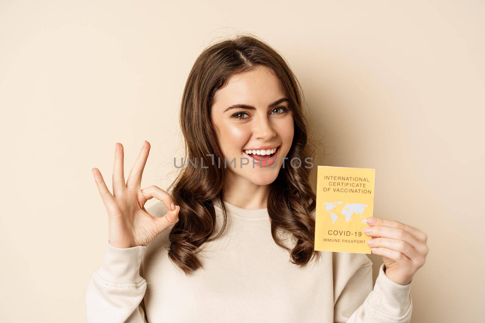 Travelling and pandemic concept. Portrait of woman showing international covid vaccination certificate, okay sign, easy travel pass, beige background.