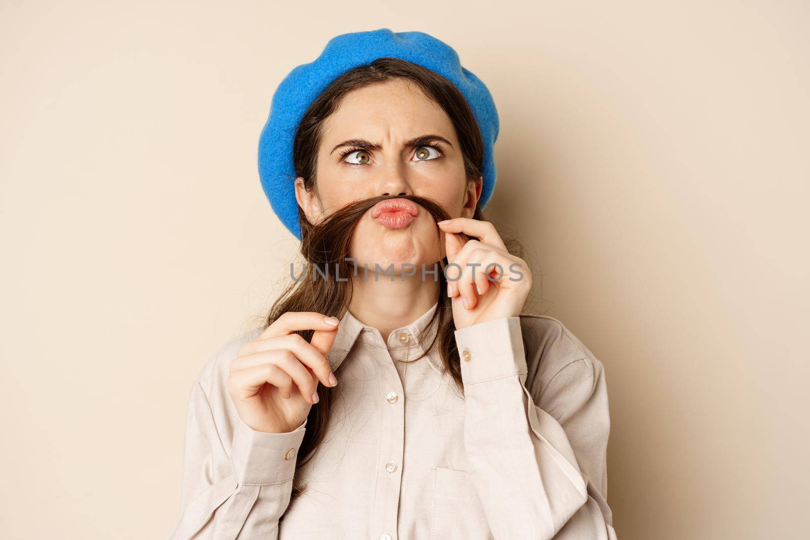 Funny beautiful girl fooling around, making moustache out of hair under lips, squinting and grimacing, standing happy against beige background.