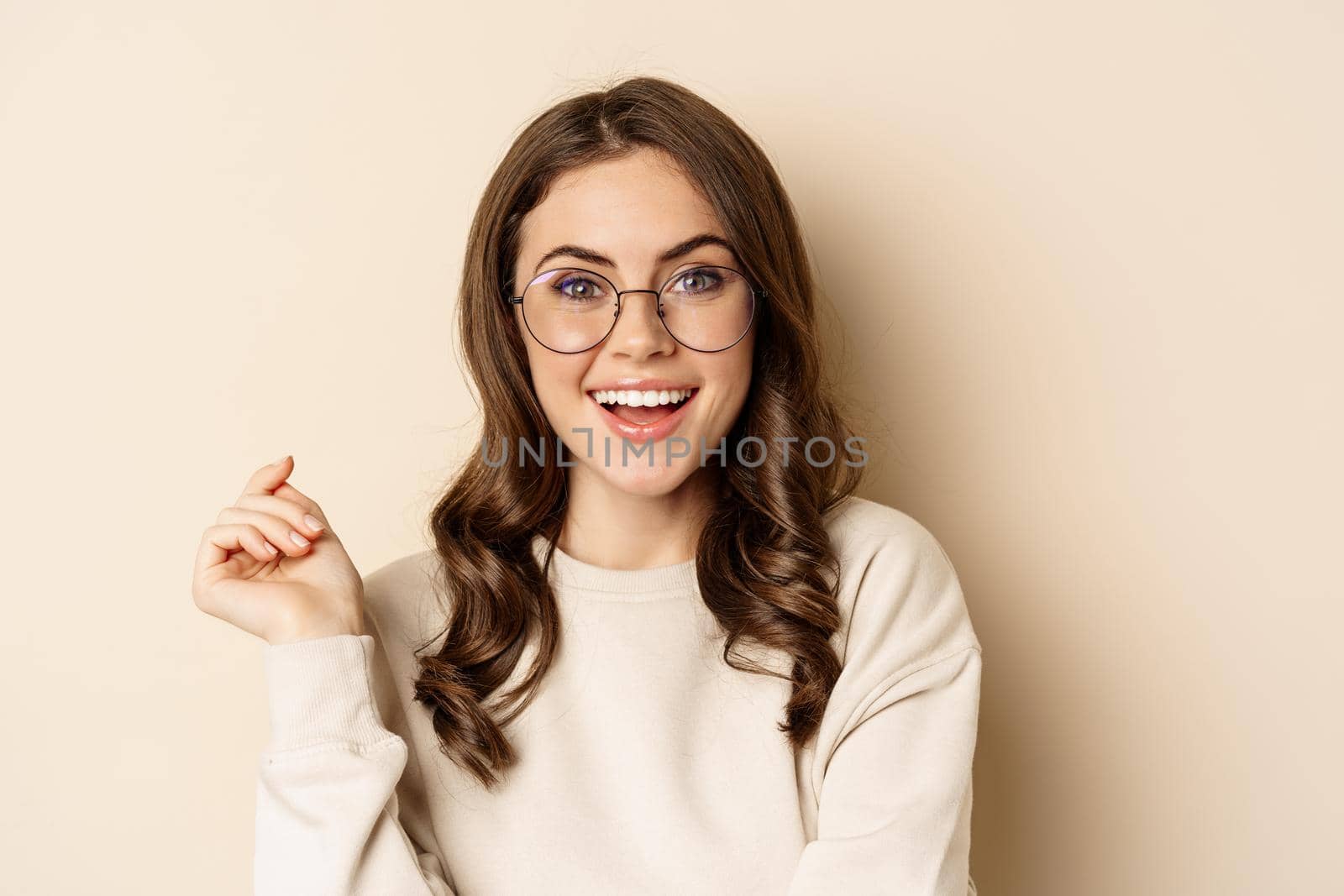 Close up portrait of stylish brunette woman in glasses, laughing and smiling, posing in eyewear against beige background by Benzoix