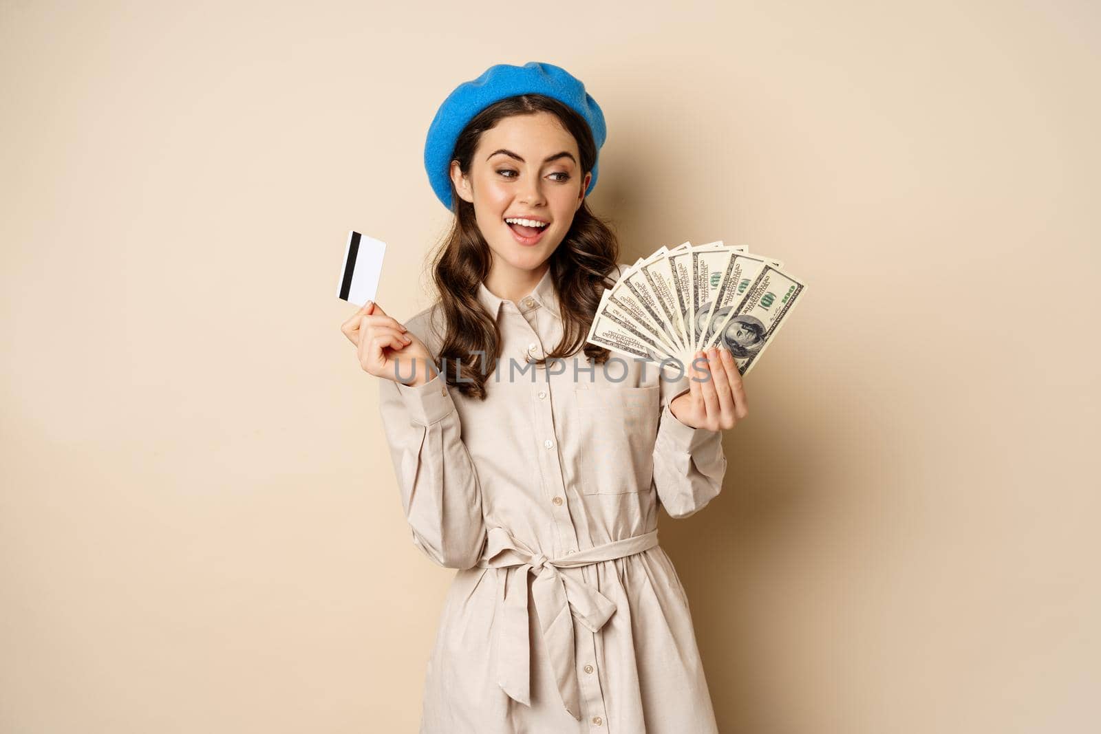 Microcredit and money concept. Young stylish woman showing credit card and dollars cash, smiling happy and satisfied, standing over beige background.