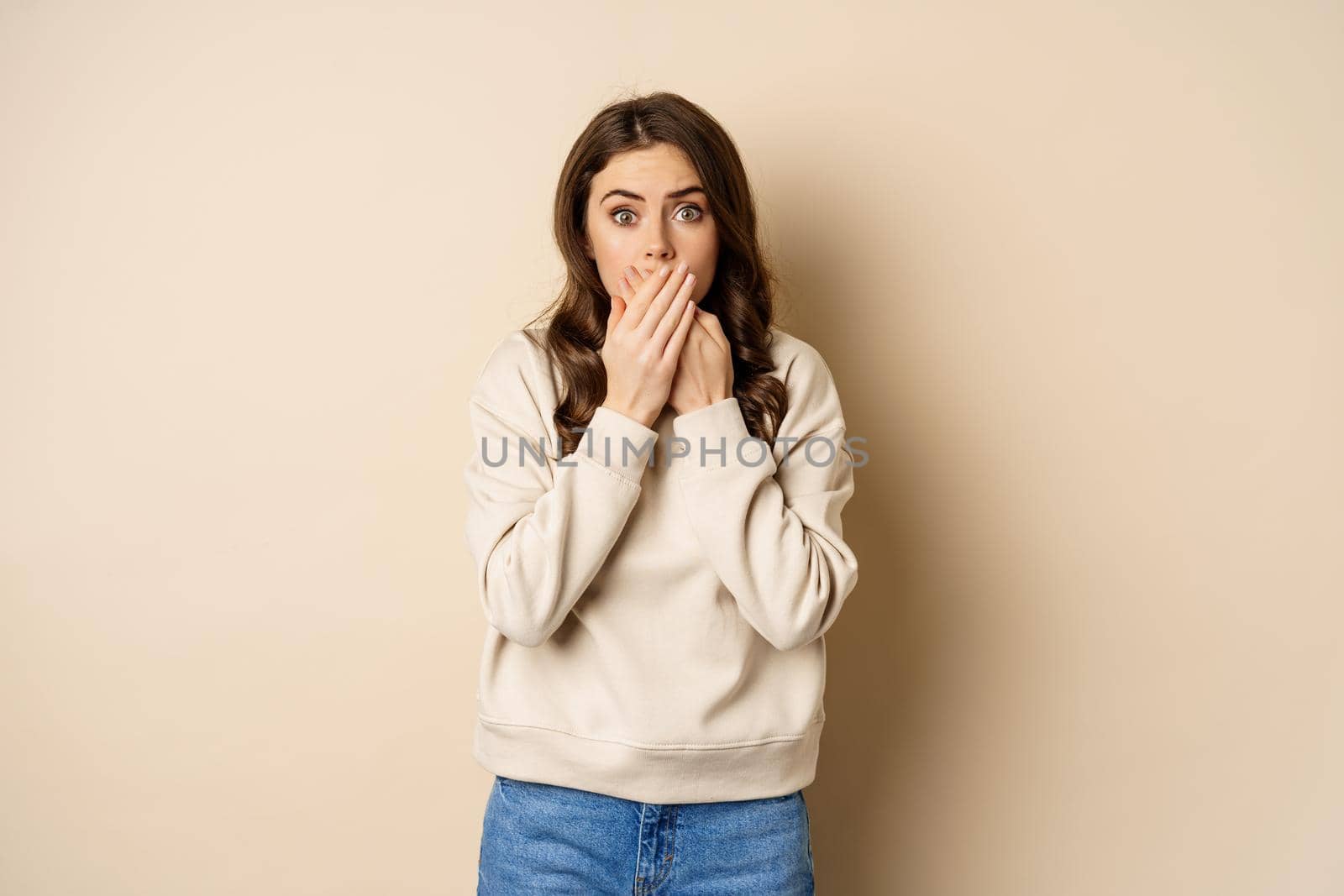 Shocked and scared woman cover mouth, looking stunned and speechless, worry about smth, standing over beige background.