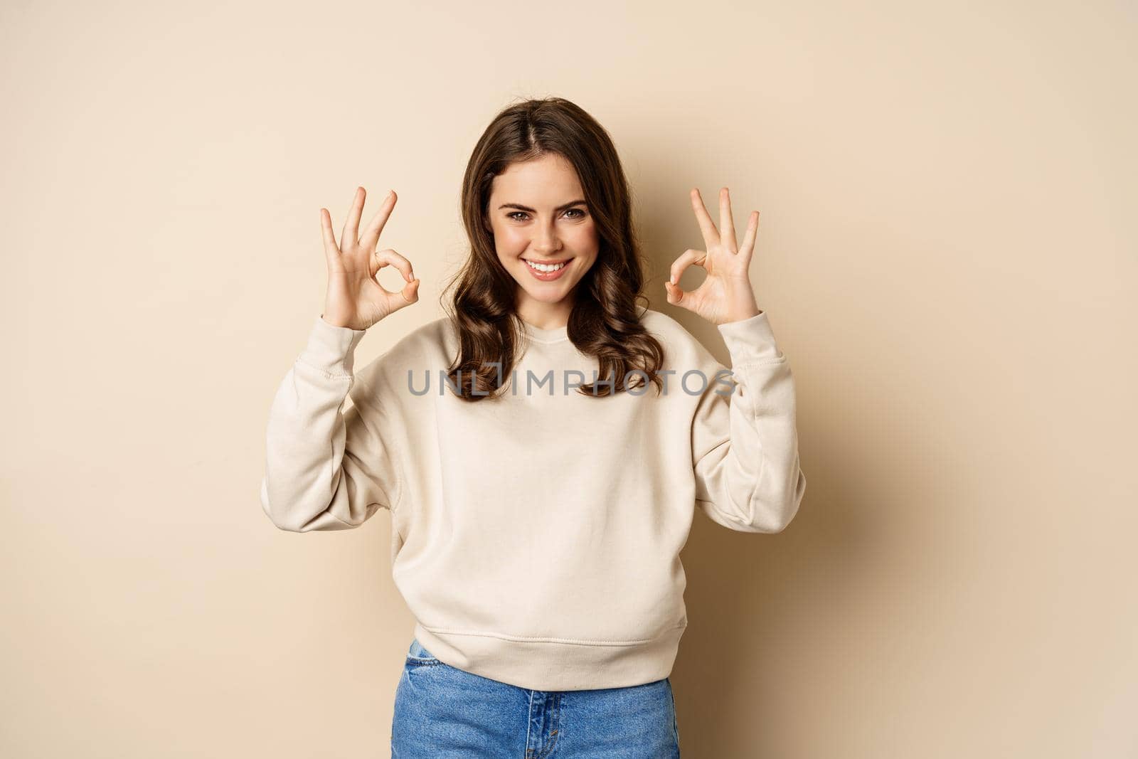 Okay. Confident smiling modern girl showing ok sign, approve, like and recommend smth good, standing over beige background.