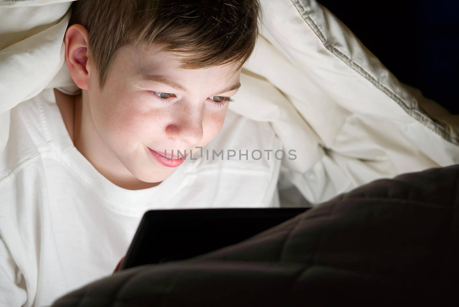 Distance learning. Online education. Happy boy in bed under blanket with tablet in dark. Child's face is illuminated by bright monitor. Quarantine. social media addiction by PhotoTime