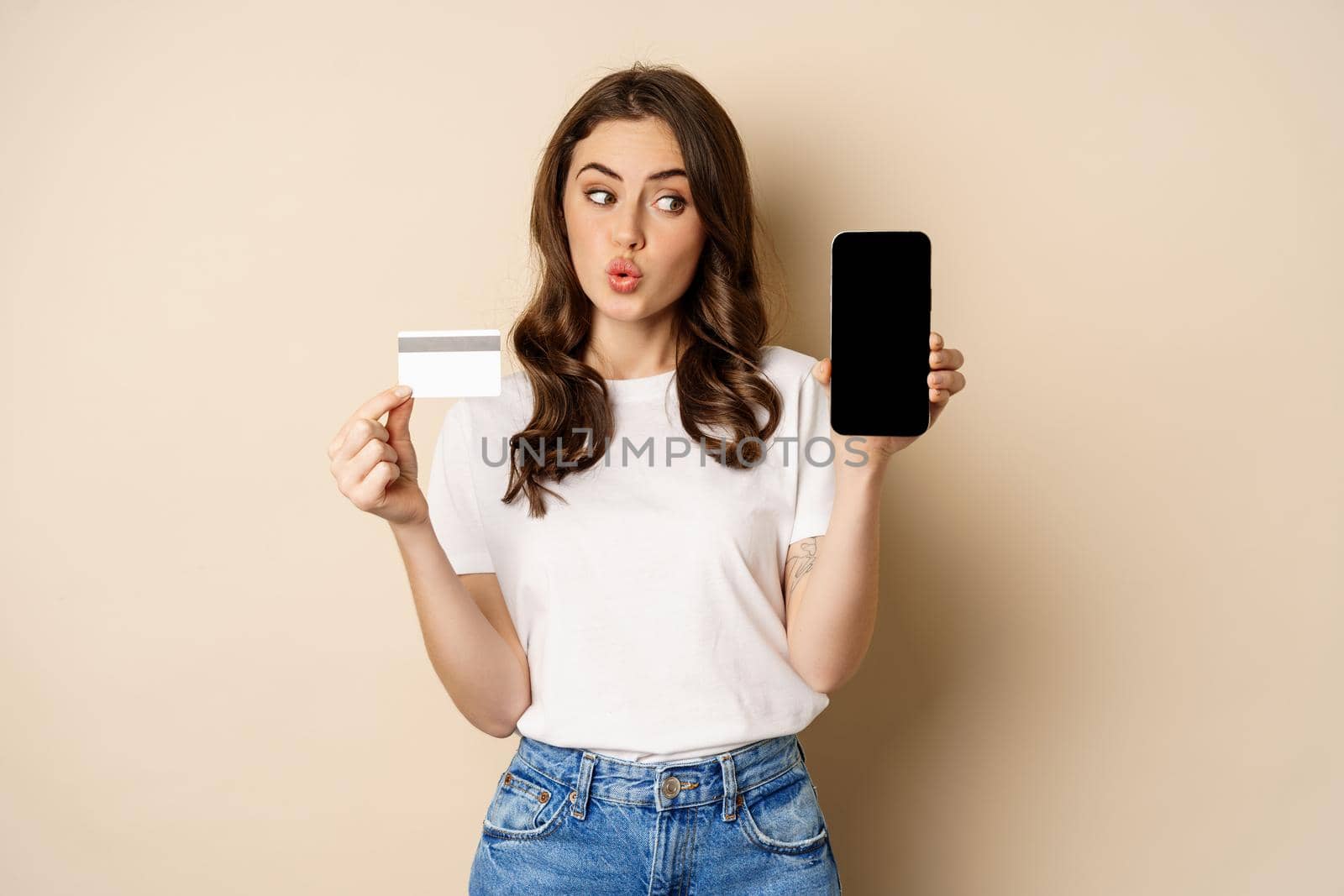 Online shopping and people concept. Young beautiful woman looking happy, showing credit card discount and mobile phone screen, standing over beige background.