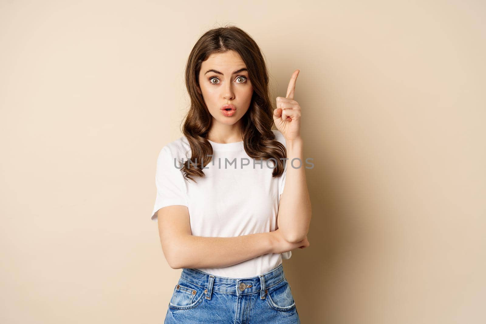 Excited woman raising finger, pitching an idea, has suggestion, wearing white t-shirt and jeans over beige background.
