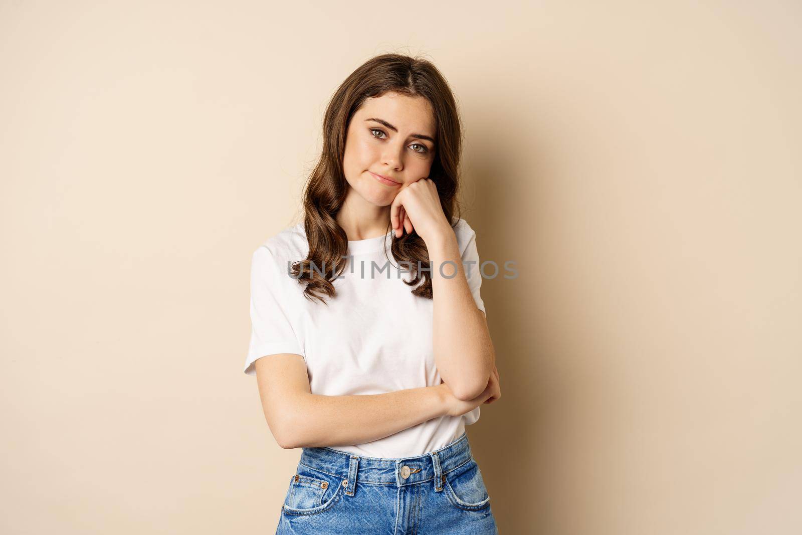 Sad and bored brunette girl looking unamused, uninterested, standing over beige background.