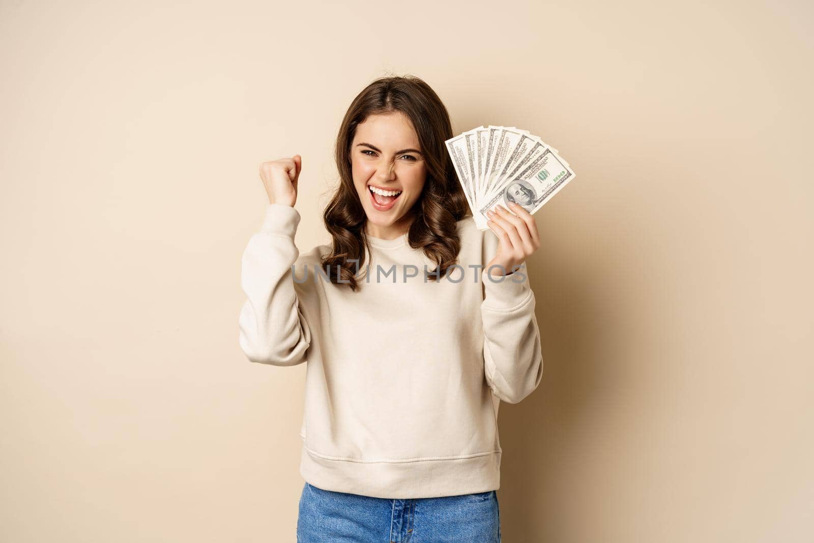 Enthusiastic modern woman winning money, got cash, celebrating and shouting of joy, standing against beige background.