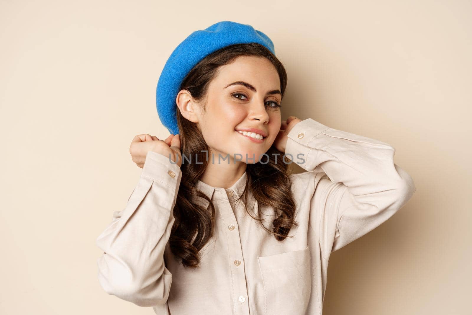 Stylish modern girl put on trendy hat on head and smiling, going out, posing against beige background.