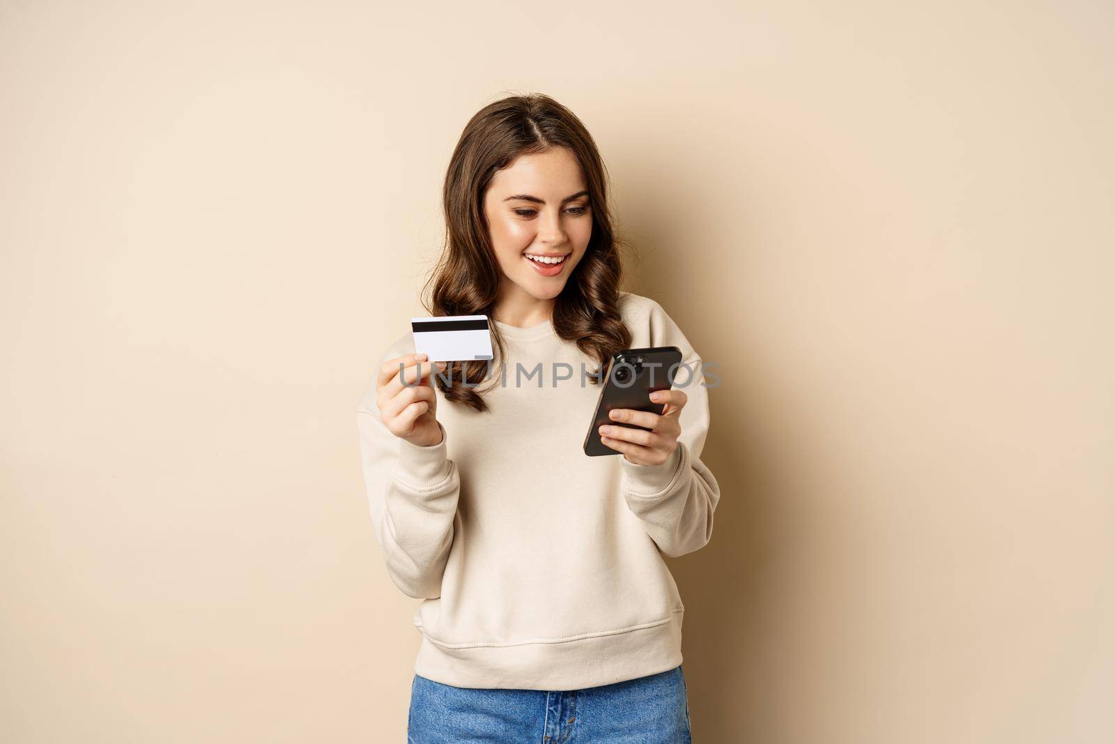 Smiling girl using mobile app, smartphone shopping and credit card, standing over beige background, order smth by Benzoix