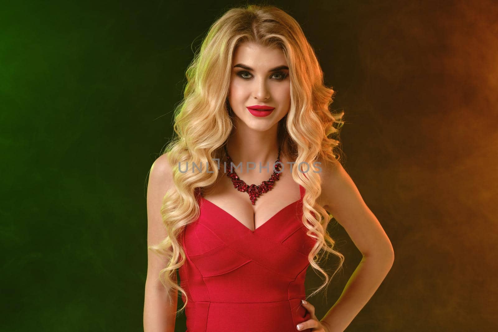 Charming curly blonde girl with bright make-up, in red fitting dress and necklace. She is smiling, hand on waist, posing on colorful smoky studio background. Fashion and beauty. Close up, copy space
