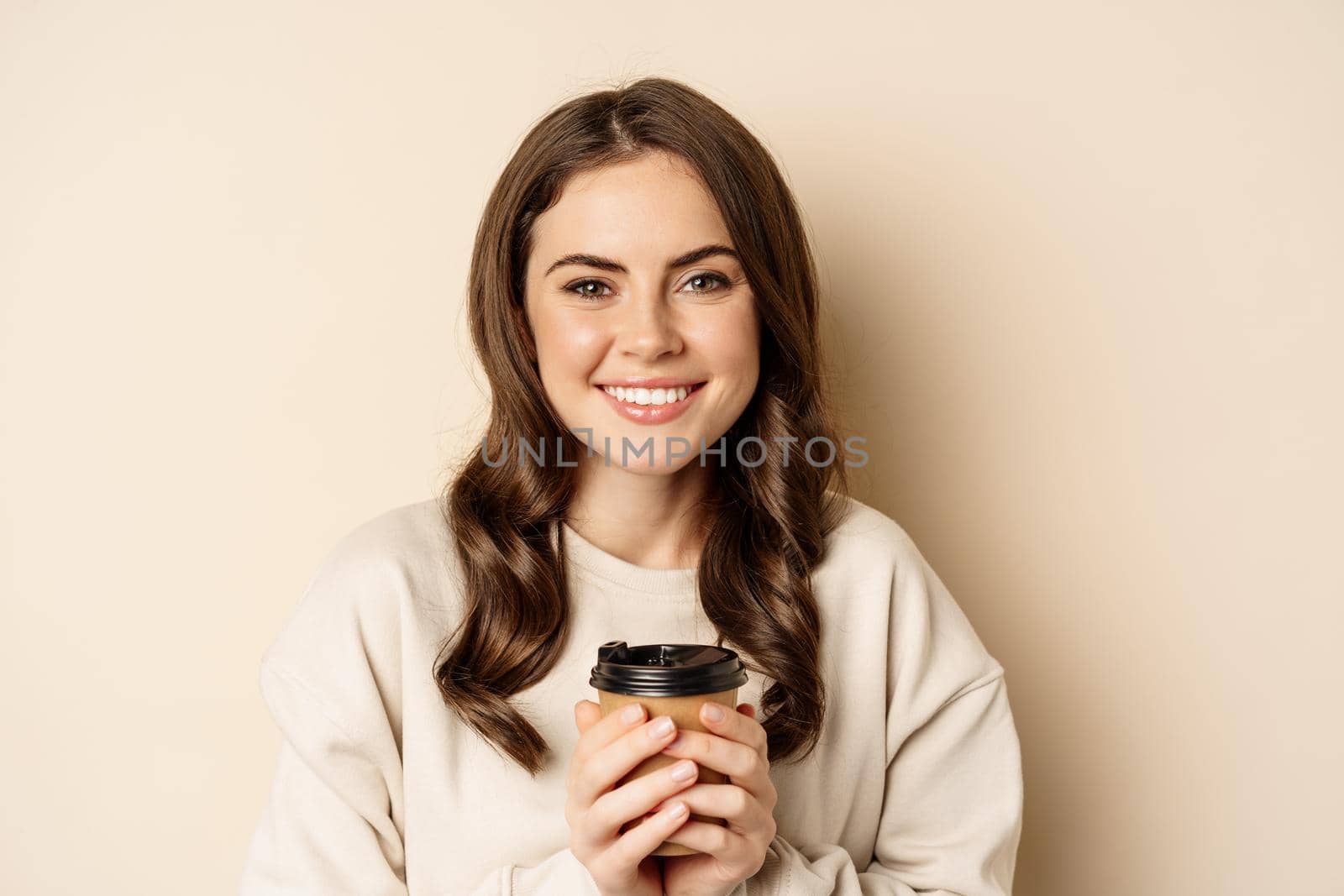 Beautiful authentic woman smiling, holding warm cup of coffee and looking happy, standing over beige background by Benzoix