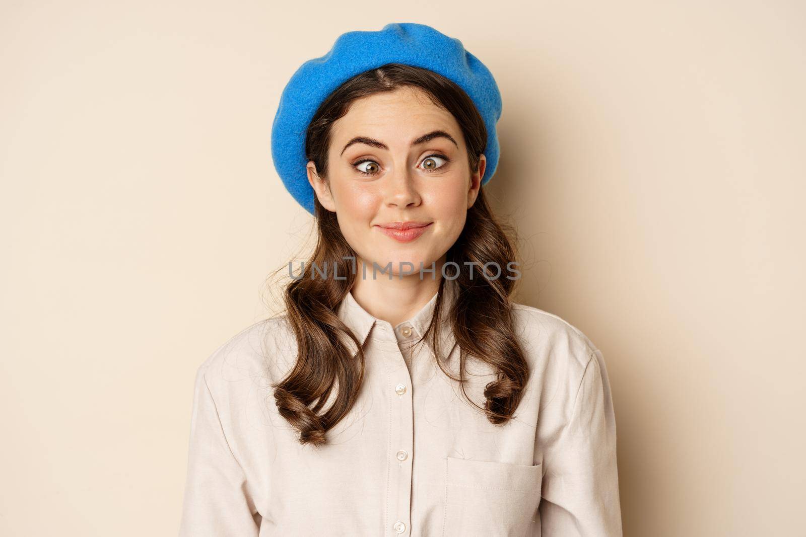 Close up portrait, silly face of young funny girl, squinting eyes and having fun, posing in trench and hat against beige background.