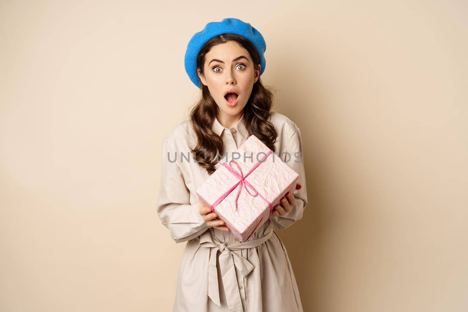 Holidays and gifts concept. Beautiful woman holding gift box with surprised, disbelief face, receive gift, beige beige background.