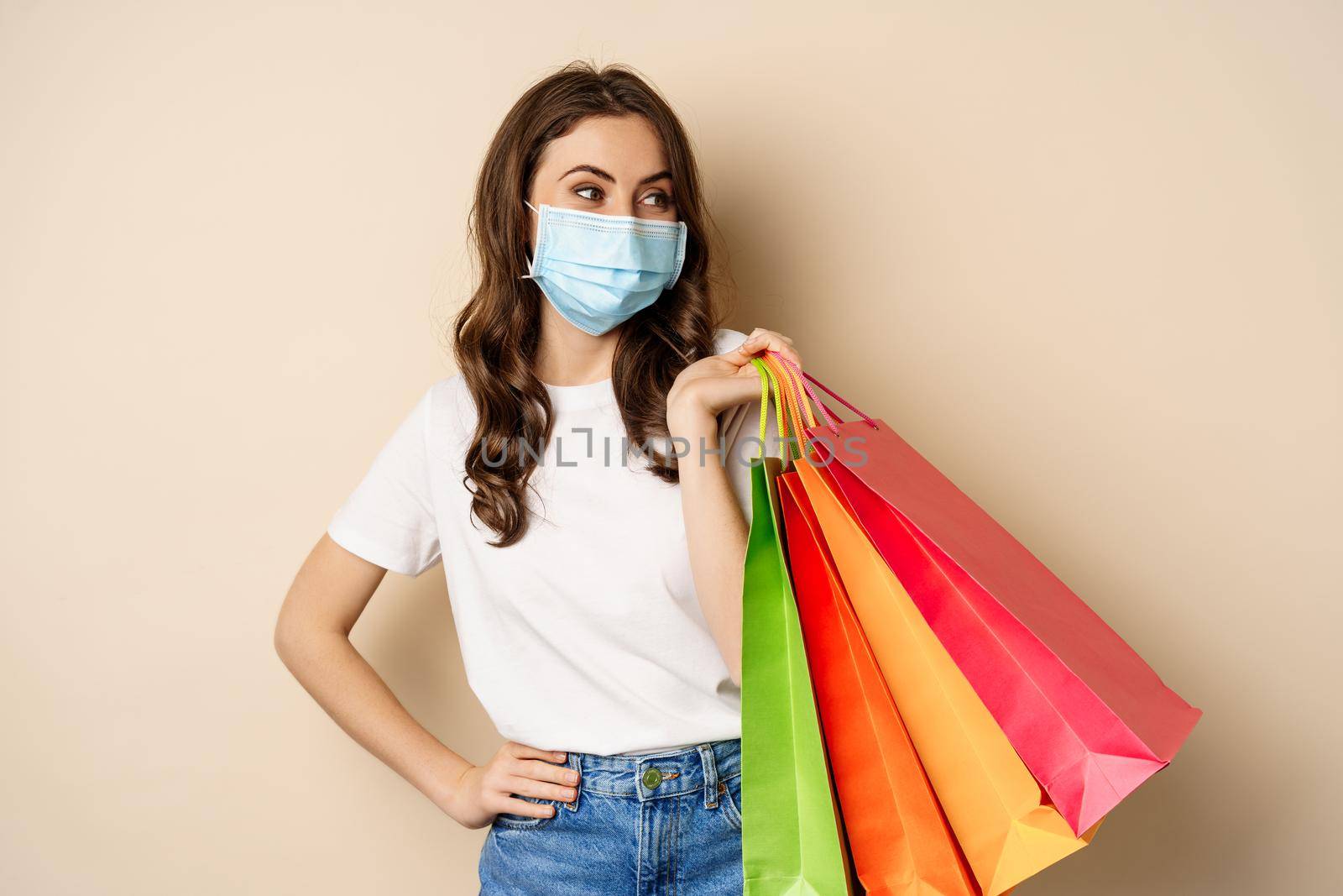 Covid-19 pandemic and lifestyle concept. Young woman posing in medical face mask with shopping bags from mall, vaccinated girl going in store in personal protective equipment, beige background by Benzoix