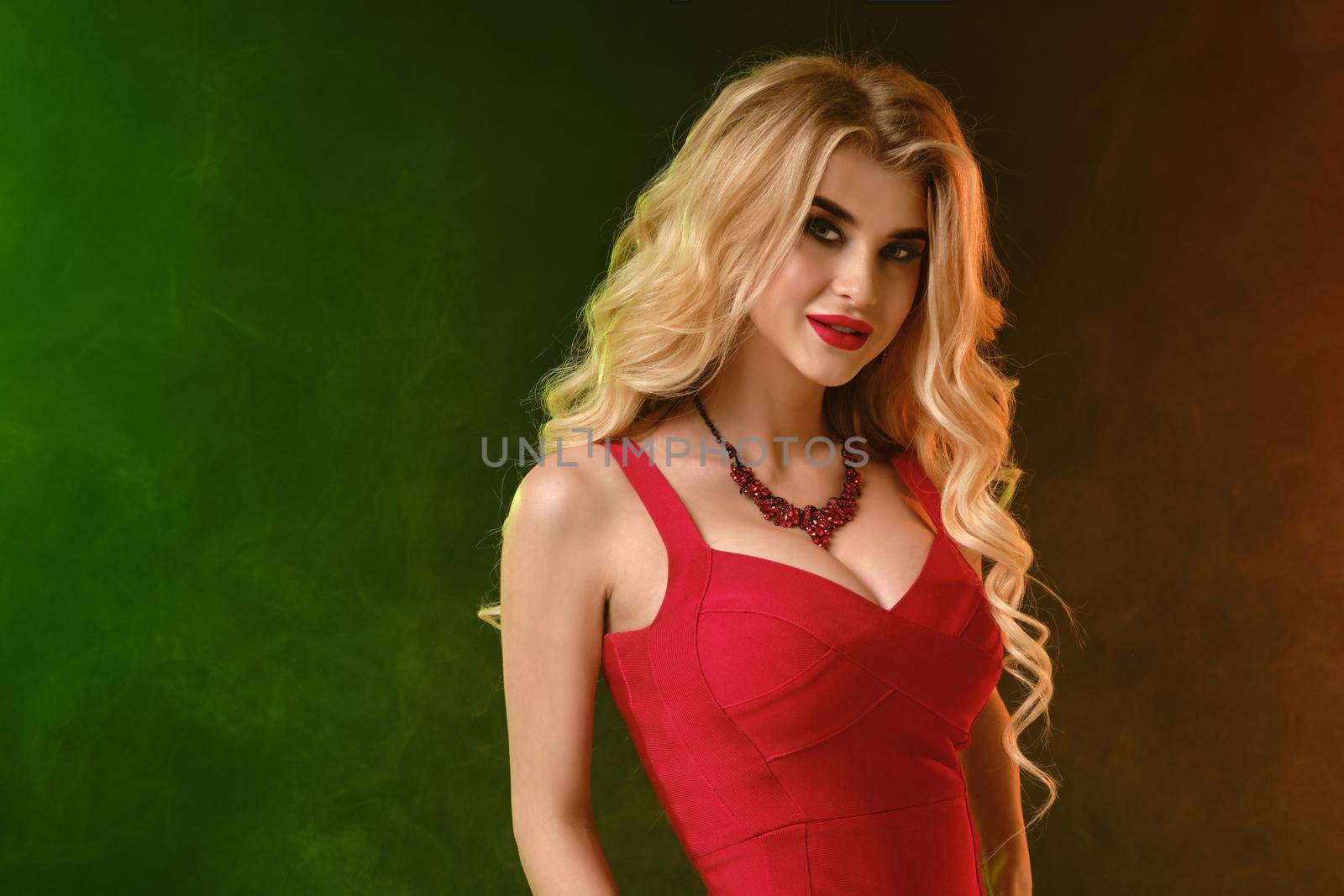 Charming curly blonde woman with bright make-up, in red fitting dress and necklace. She is smiling while posing against colorful smoky studio background. Fashion and beauty. Close up, copy space