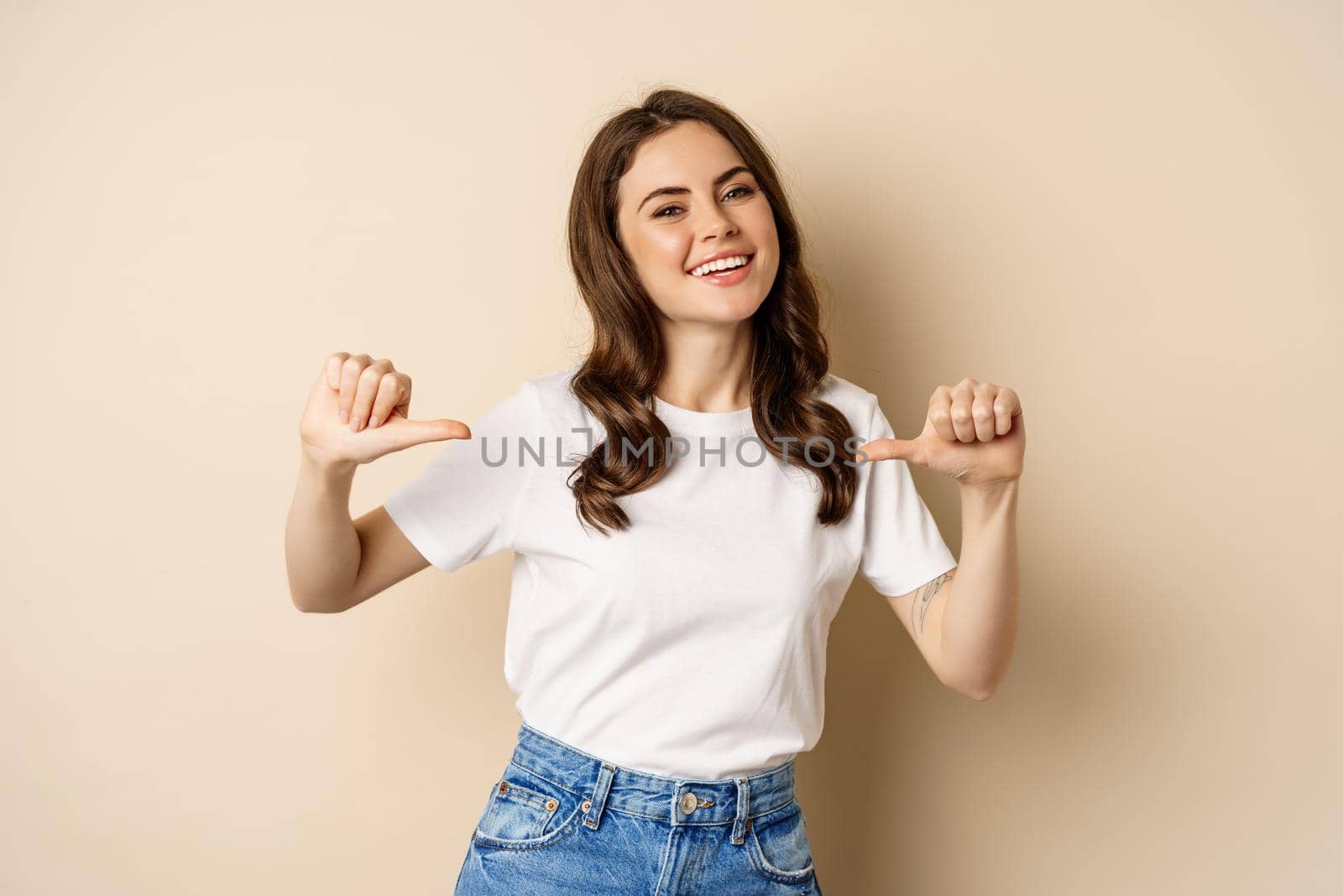 Self-assured young happy woman pointing fingers at herself and dancing, self-promoting, being confident, standing over beige background.